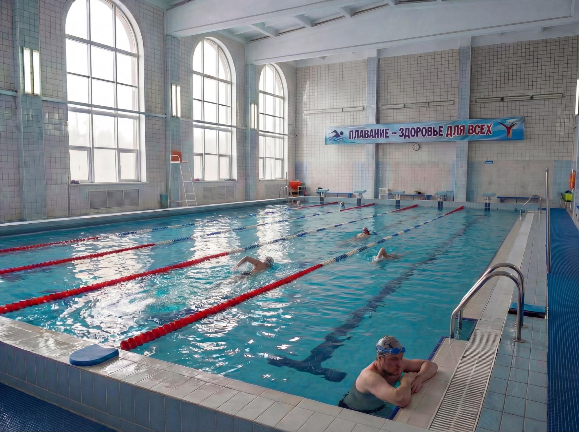 Крытый плавательный бассейн с несколькими дорожками, разделёнными красными буйками. Несколько человек плавают, один отдыхает у бортика. На стене баннер с надписью «Плавание — здоровье для всех» Крытый плавательный бассейн с несколькими дорожками, разделёнными красными буйками. Несколько человек плавают, один отдыхает у бортика. На стене баннер с надписью «Плавание — здоровье для всех»