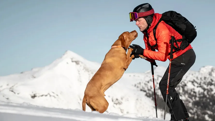 Мужчина в экипировке и с лыжами компании ATOMIC играет с собакой.