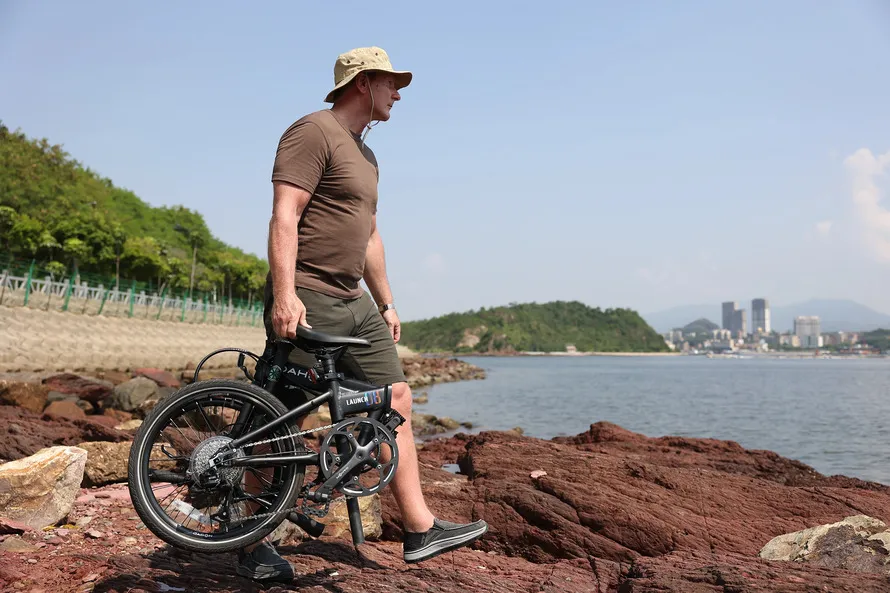 Мужчина идет к реке, держа в правой руке сложенный велосипед производства компании DAHON.