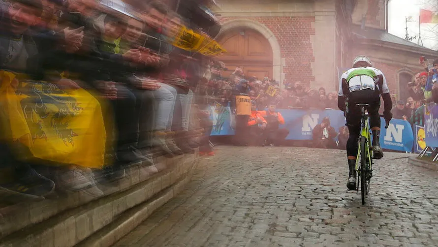 Велосипедист едет на велосипеде по брусчатке между зрителями.