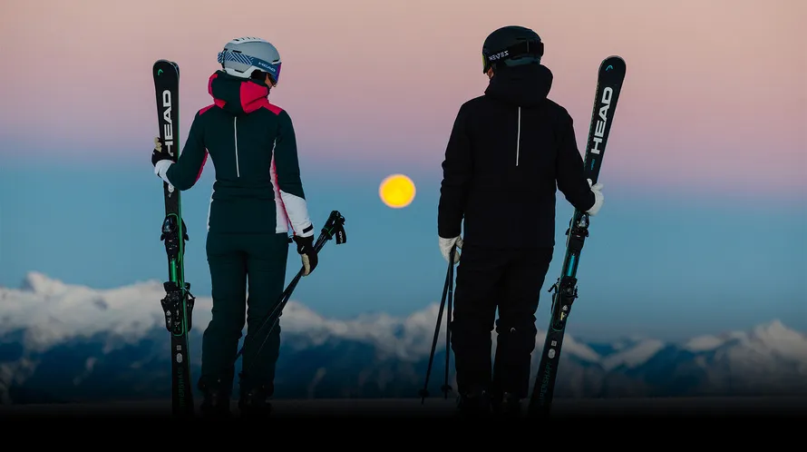 Мужчина и женщина с горными лыжами и в экипировке бренда HEAD смотрят на закат.