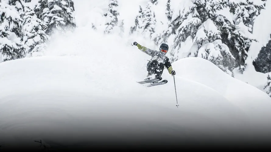 Фрирайдер подпрыгивает на заснеженном холме, мчась вниз с горы на горных лыжах с креплениями производства компании TYROLI.