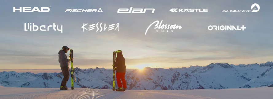 Мужчина и женщина с горными лыжами стоят на снежной трассе на фоне заката и логотипов партнеров компании TYROLIA.